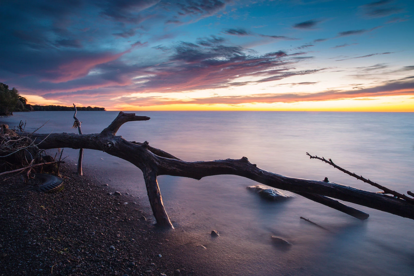 Driftwood stretches from a pebbled shoreline into calm water under a colorful evening sky