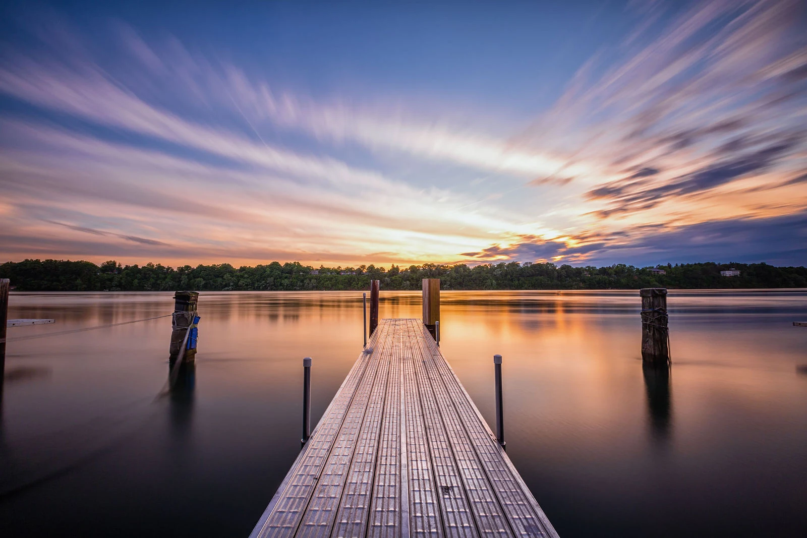 A wooden pier leads across still water toward a glowing sunset and streaked clouds